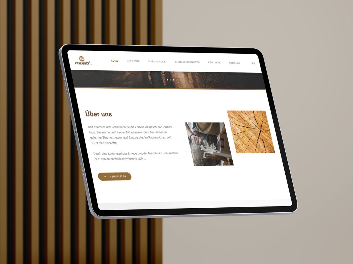 iPad in front of wooden lamellae background showing the "About us" section of the Holzbau Heidesch website.